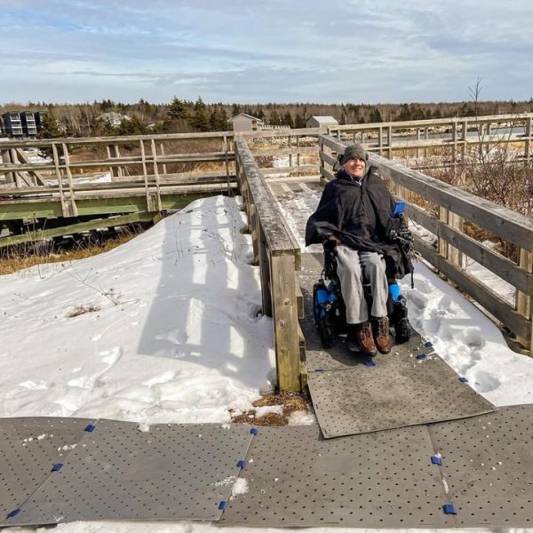 Photo shows a man smiling while seated in his power wheelchair on a grey portable access pathway over snow on a small wooden bridge outdoors. He is wearing winter clothes and a beanie.