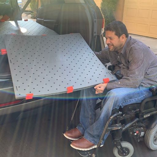 A man smiles while holding a stack of square grey access mats (Access Trax) half way in the trunk of his accessible van. He is seated in his power wheelchair and is wearing a long sleeve button up shirt and jeans with brown shoes.