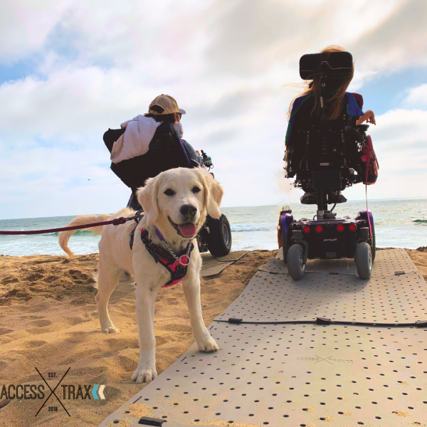 Photo of a man and woman seated in their power wheelchairs at the beach facing away from the camera to the ocean. Their chairs are on a grey portable access mat called Access Trax. There is a white lab dog smiling behind them.