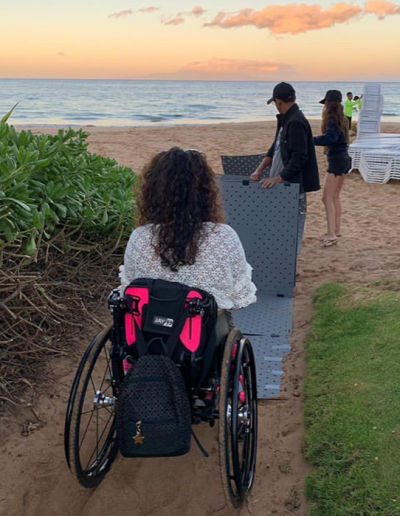 A woman named Gretchen watches two people unfold Access Trax mats onto sand at a Maui beach during sunset. She is seated in her manual wheelchair and has a black backpack hanging from her chair. She has long brown curly hair and is wearing a white blouse.