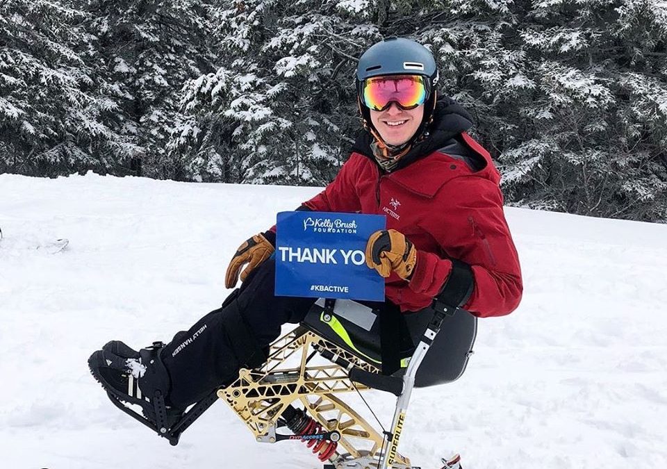 A man smiles while seated in his monoski in a snowy mountaintop. He has a red snow jacket, a helmet, and goggles. He is holding a blue sign that says "Thank you" to the Kelly Brush Foundation.