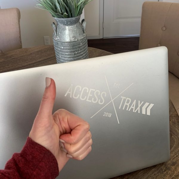 Image shows a silver lap top on a table with a white Access Trax logo sticker on it. There is a person showing "thumbs up" next to the sticker.