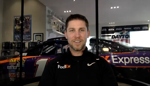 Denny Hamlin (man with light brown hair and a short beard) smiles during an interview. He is wearing a black zip up jacket with the FedEx and Nike logos and behind him is his #11 NASCAR racecar in a garage.