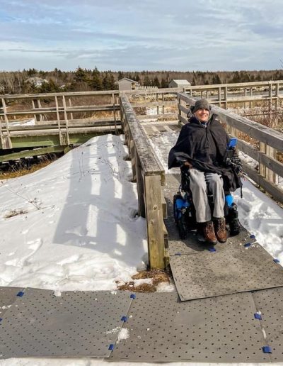 A man wearing winter clothing smiles seated in his power wheelchair on a snow covered wooden bridge. His is on grey Access Trax portable mats.