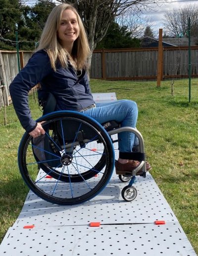 A blonde woman wearing jeans and a blue zip up jacket smiles in her grassy backyard. She is seated in her manual wheelchair which is on a grey Access Trax portable pathway.