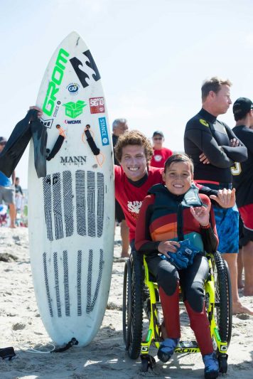 A young male adaptive surfer smiles with a surf volunteer at the beach during an adaptive surf event. He is seated in his manual wheelchair and holding up the surf "shaka" symbol with his left hand. His adaptive surf board is on their right.