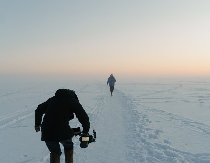 A person crouches low to the ground over snow filming another person running ahead. The sky is at dusk.