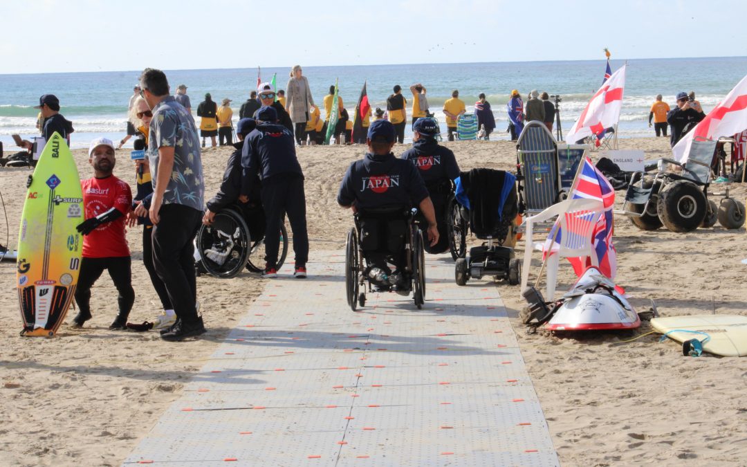 Photo of adaptive surfers at the World Para Surfing Championships on the beach. There is a portable beach access pathway people in wheelchairs are using to get to the event. In the distance, there are people near the water.