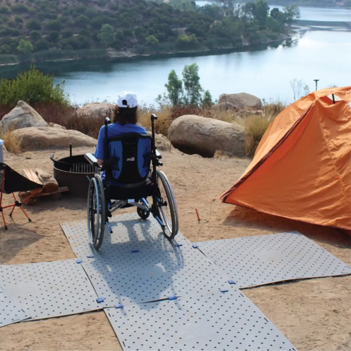 A girl in a wheelchair next to an orange tent watching a lake 