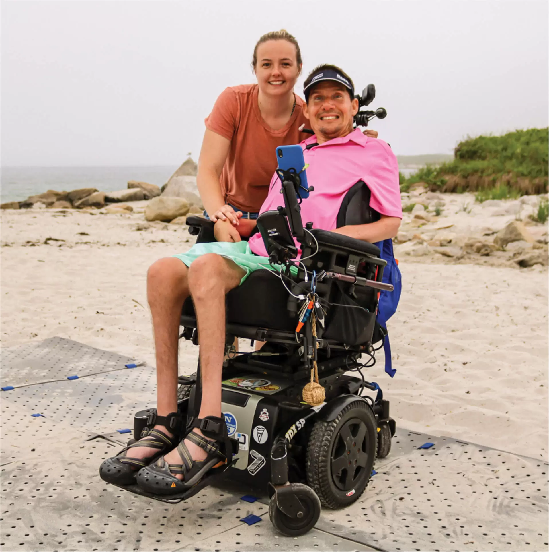 Man in a wheelchair with a pink shirt on the beach with a woman standing besides him