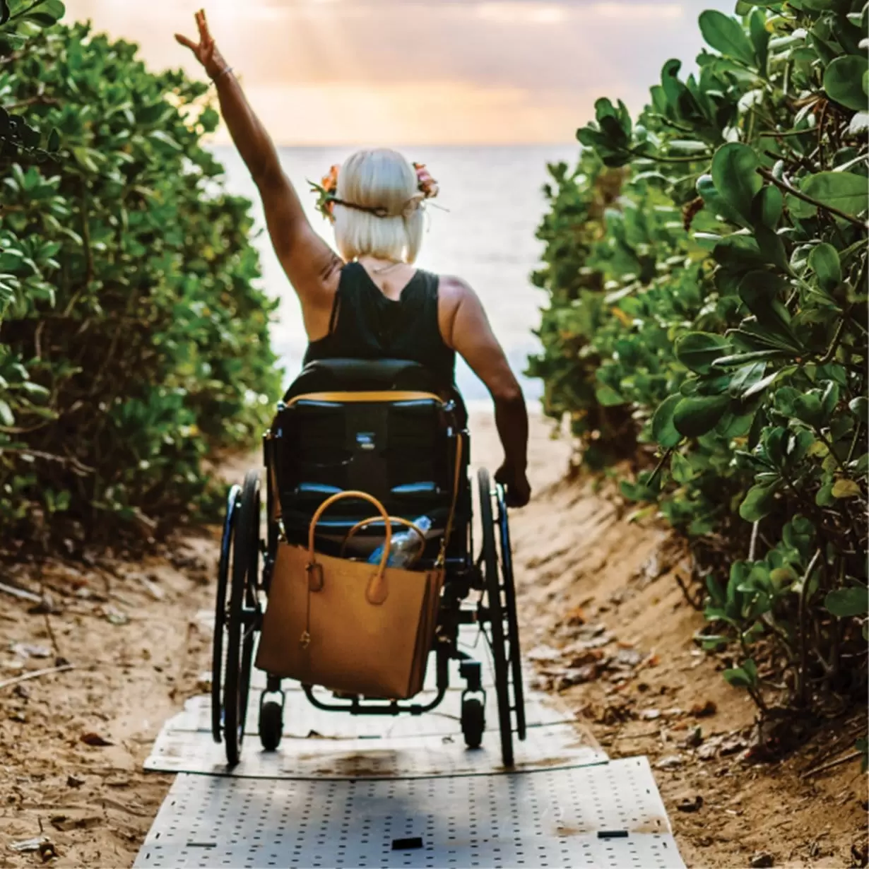 A woman in a wheelchair with a raised hand going down a road surrounded by green plants